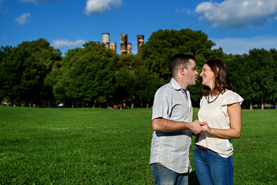 An engaged couple hold hands in penn treaty park with the smoke stacks peaking over the trees during their fishtown engagement session.